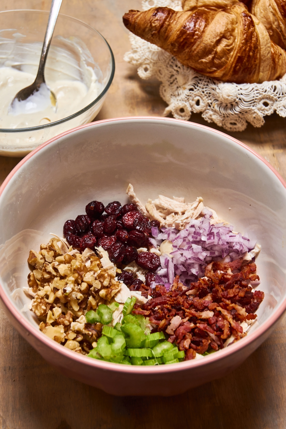 A gray bowl holds dried cranberries, red onion, nuts, celery, bacon, and chicken. A bowl full of dresssing and a croissant is near by.