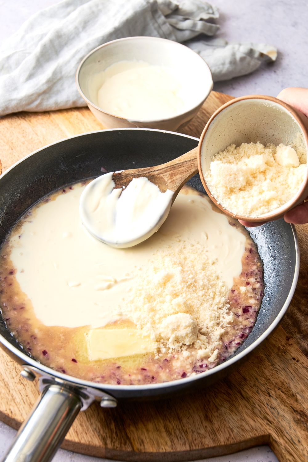 Someone dumps parmesan cheese from a small bowl into a black pan full Alfredo sauce.