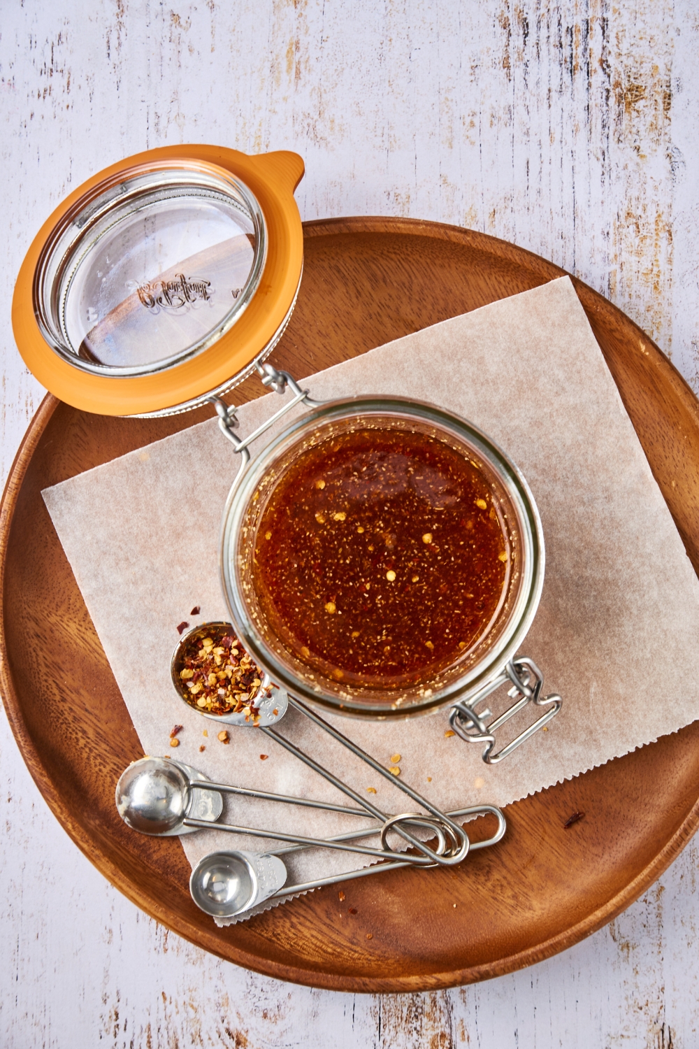 A jar full of Popeye's sweet heat sauce sits on a tray. The jar is open. A set of measuring spoons sits next to the jar. Crushed red pepper flakes are in one of the spoons.