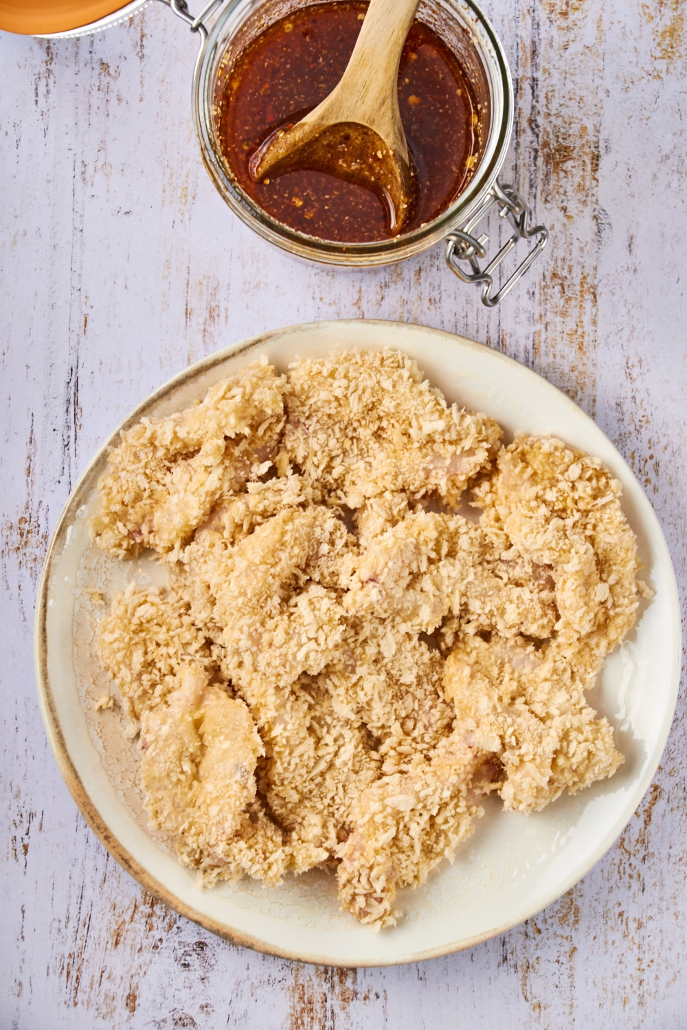 Breaded butterfly shrimp are on a plate ready to be fried. A jar of hot sauce is near by. A wooden spoon is resting in the jar of hot sauce.