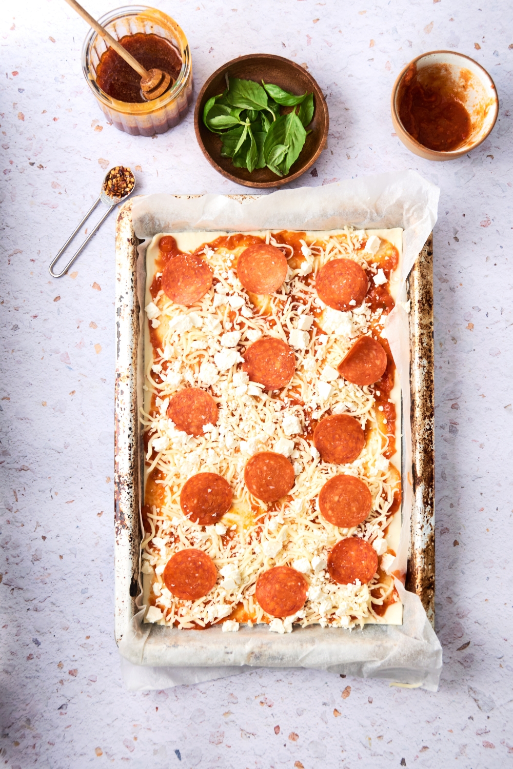 A parchment paper lined baking tray holds a pizza crust topped with pepperoni and cheese.