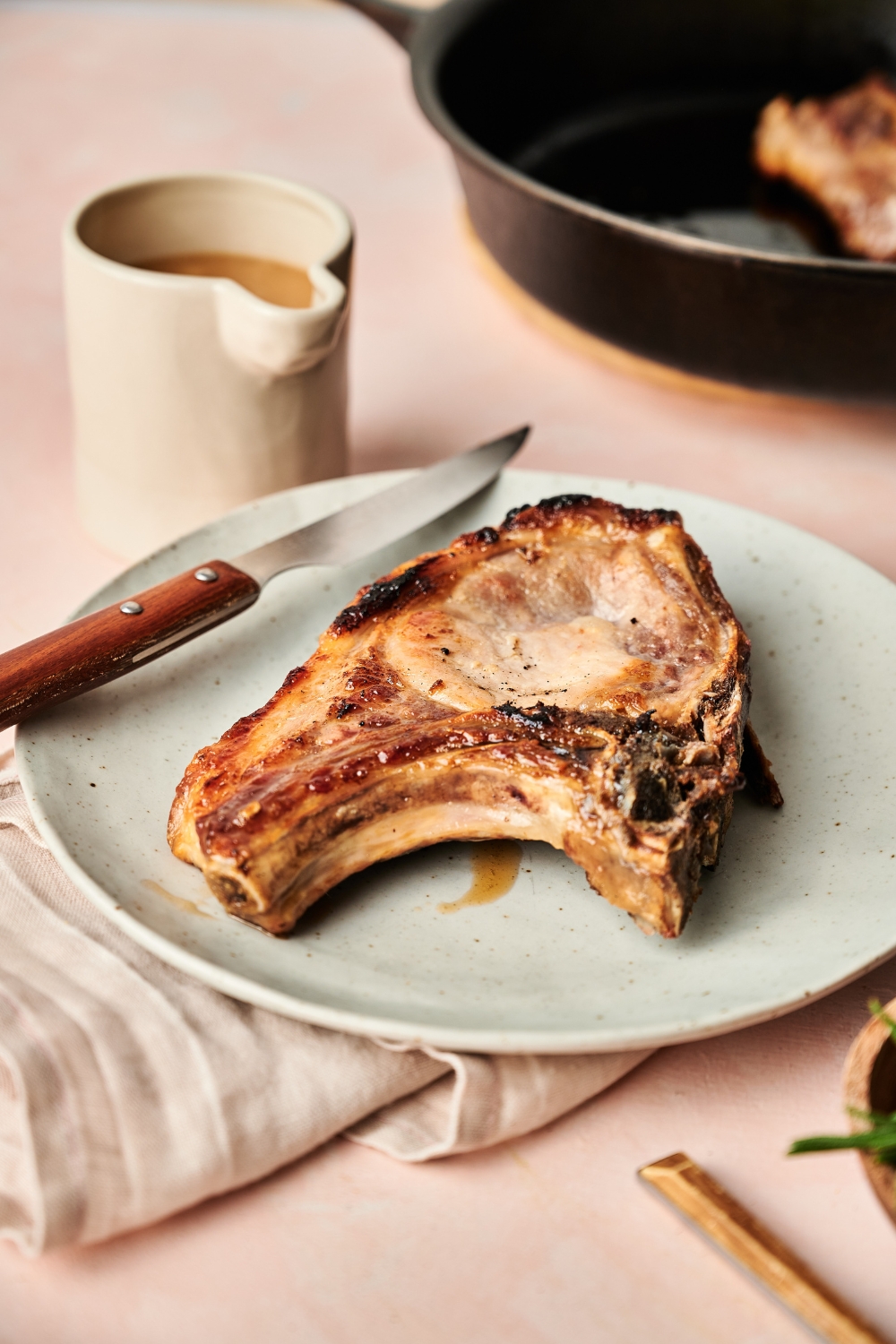 A pork chop rests on a gray plate. A knife is resting on the plate as well. A jar of gravy is near by.