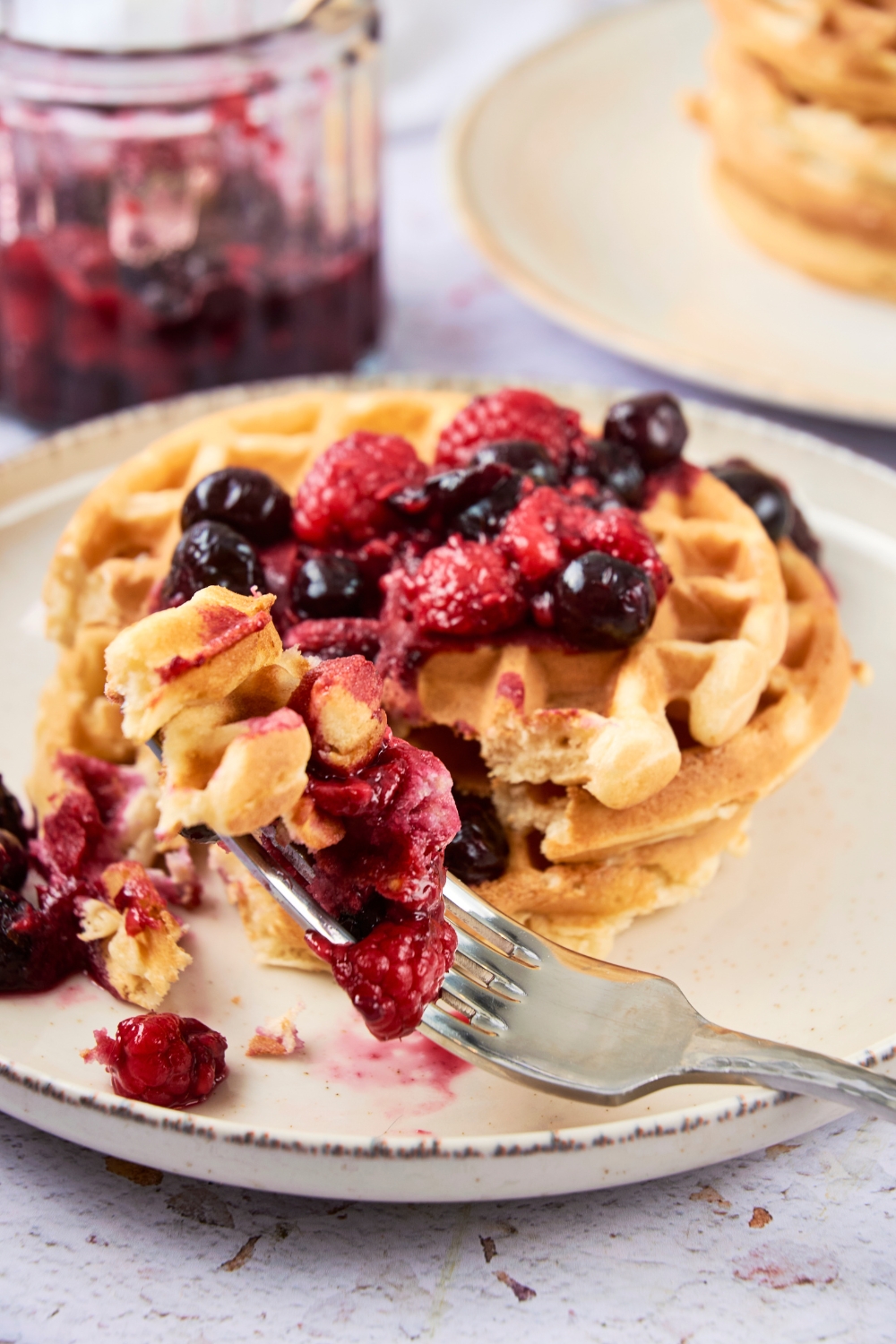 A forkful of waffles and berries rests on a plate full of waffles. A jar of berries is in the background.