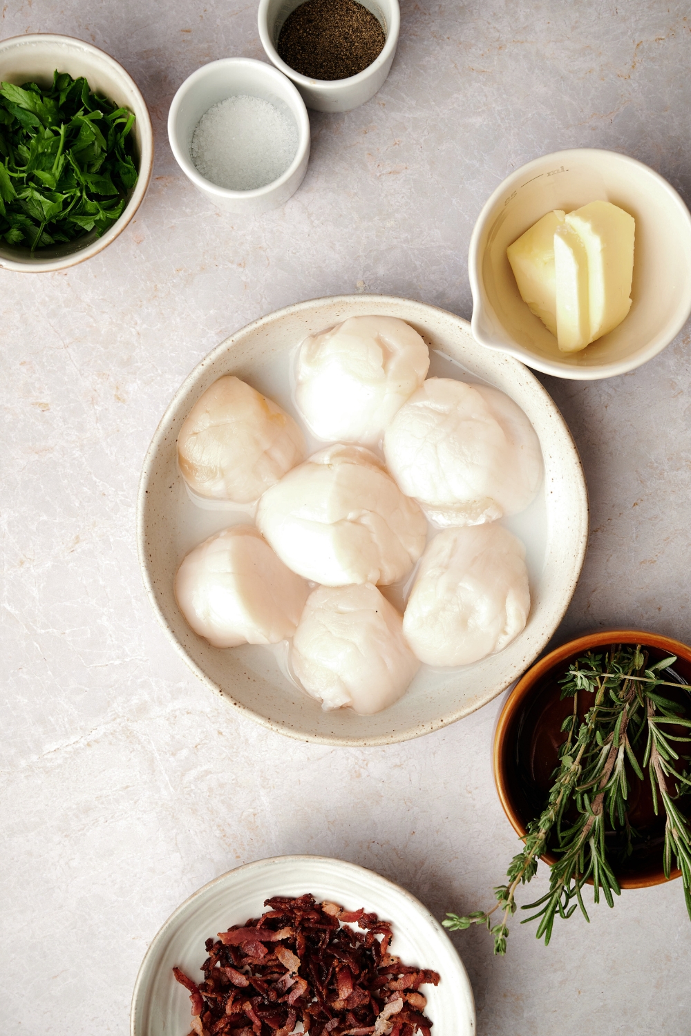 A countertop with scallops, butter, salt, pepper, parsley, and bacon bits in various bowls.