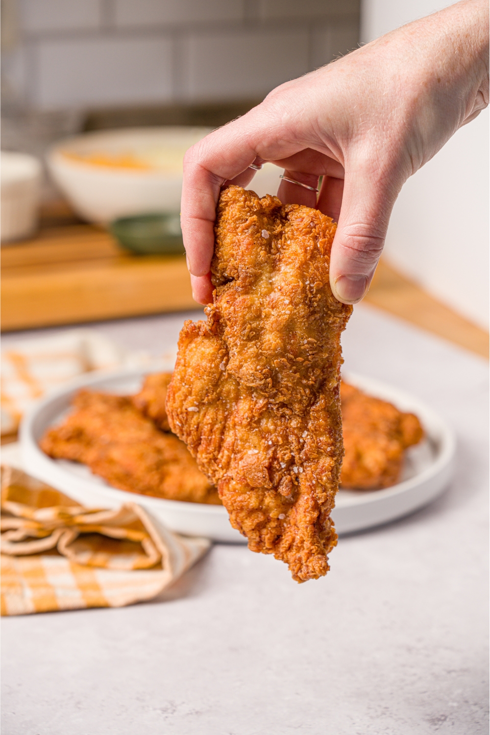 A hand holding a piece of fried chicken breast. Behind it is a white plate with a bunch of fried chicken breast on it.