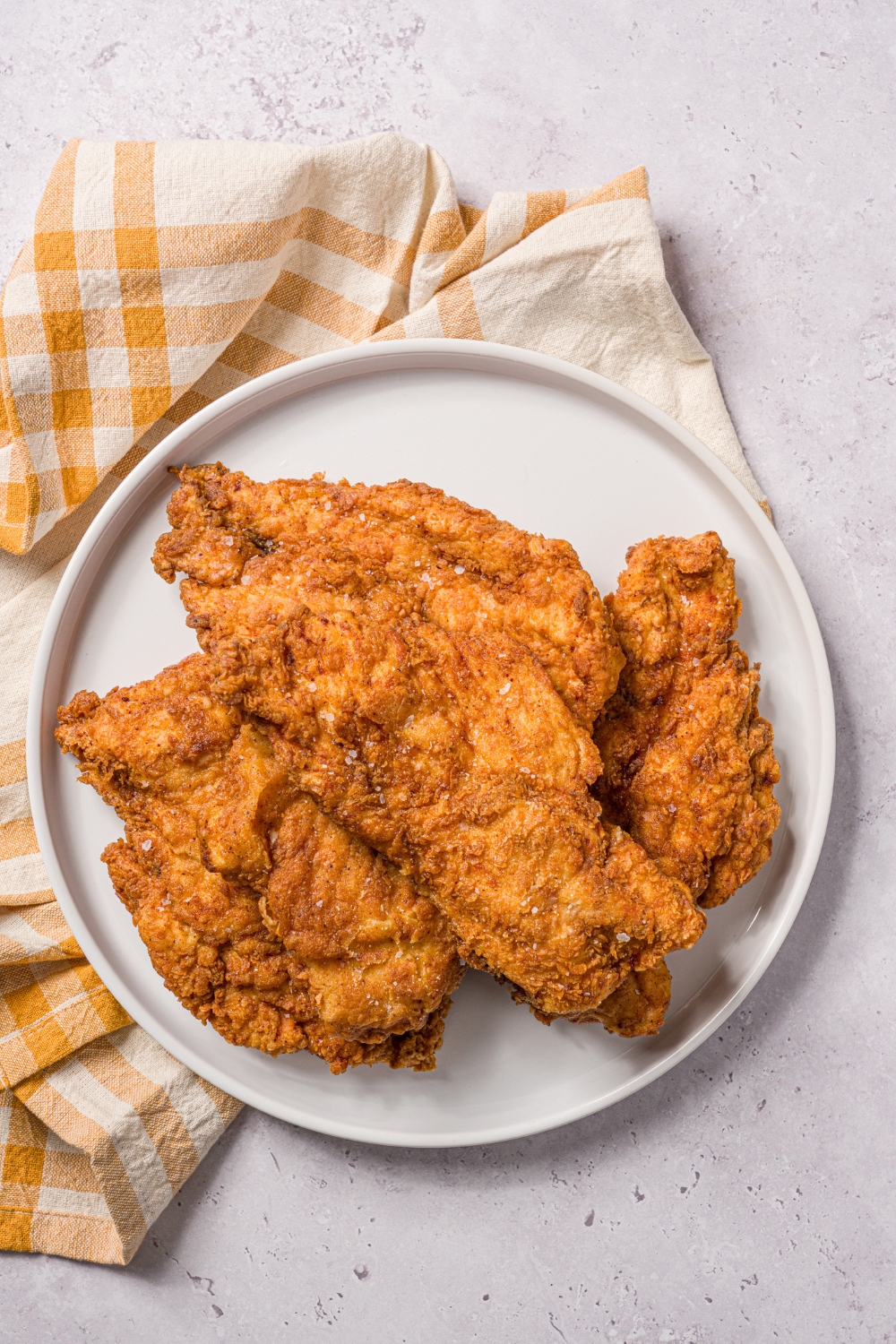 A bunch of pieces of fried chicken breast on a white plate on top of a yellow and white plaid tablecloth on a white counter.
