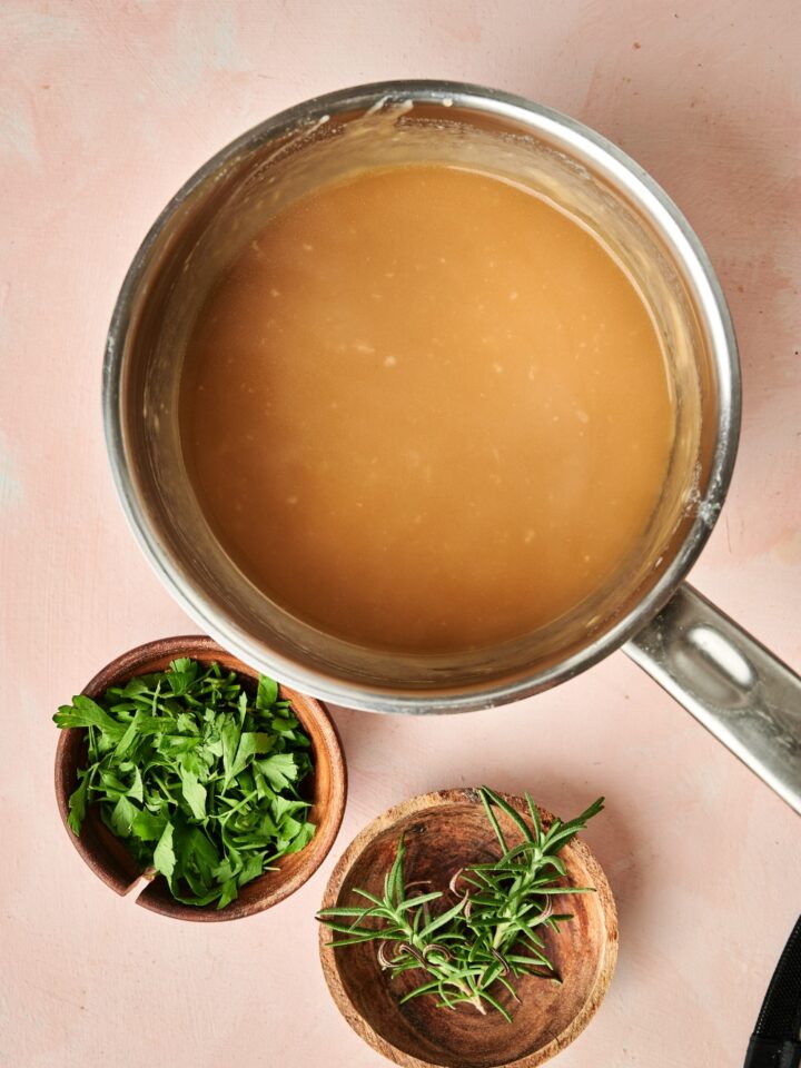 A sauce pan is full of pork gravy. It sits on the counter next to wooden bowls full of fresh herbs.