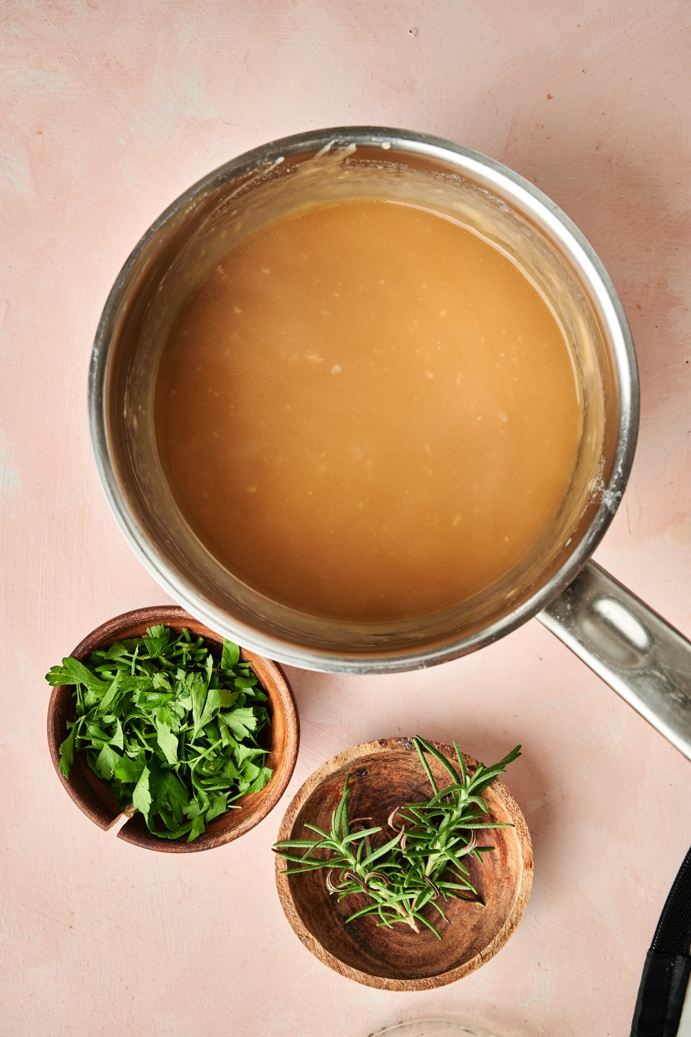 A sauce pan is full of pork gravy. It sits on the counter next to wooden bowls full of fresh herbs.