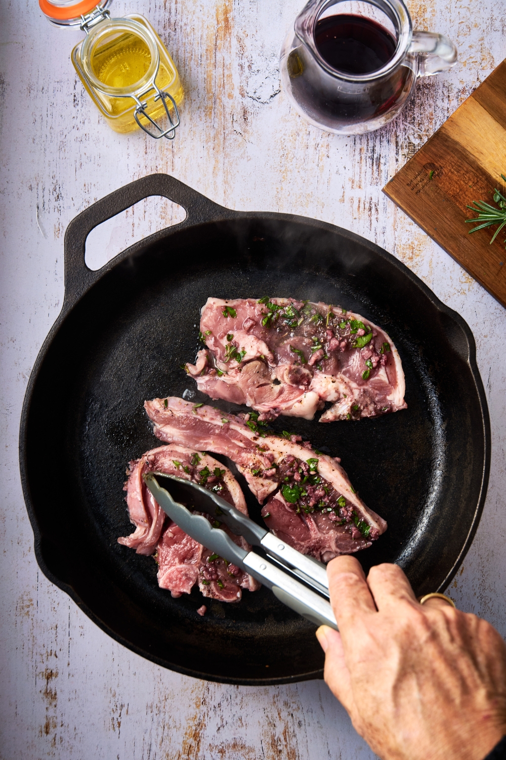 Someone presses lamb shoulder chops into a black cast iron pan. The chops are garnished with fresh parsley.