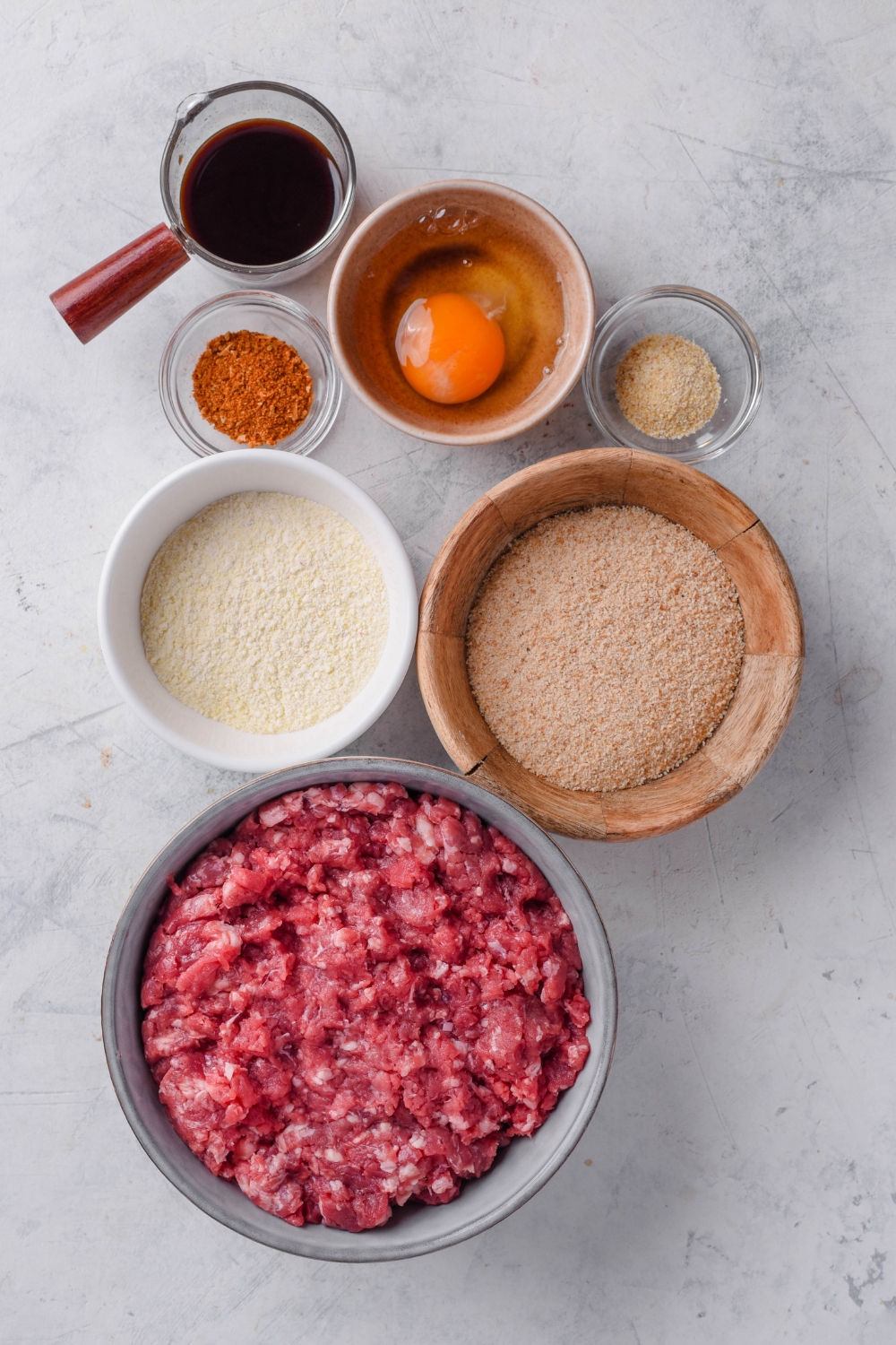 A countertop with bread crumbs, seasonings, egg, sauce, and ground beef.
