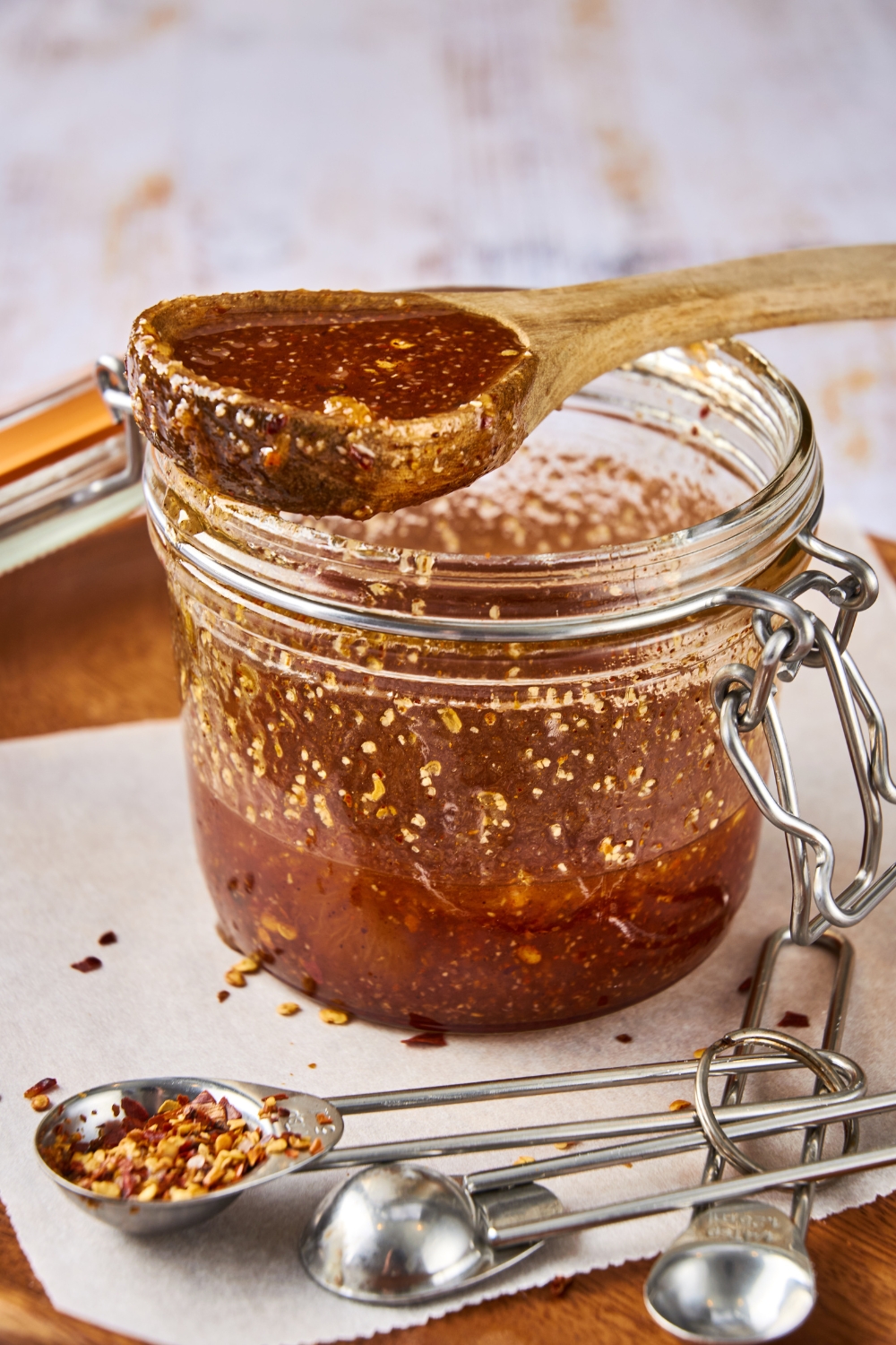 A glass jar full of Popeye's sweet heat sauce sits on a counter top. A wooden spoon covered in sauce rests on the open jar. There are measuring spoons next to the jar.