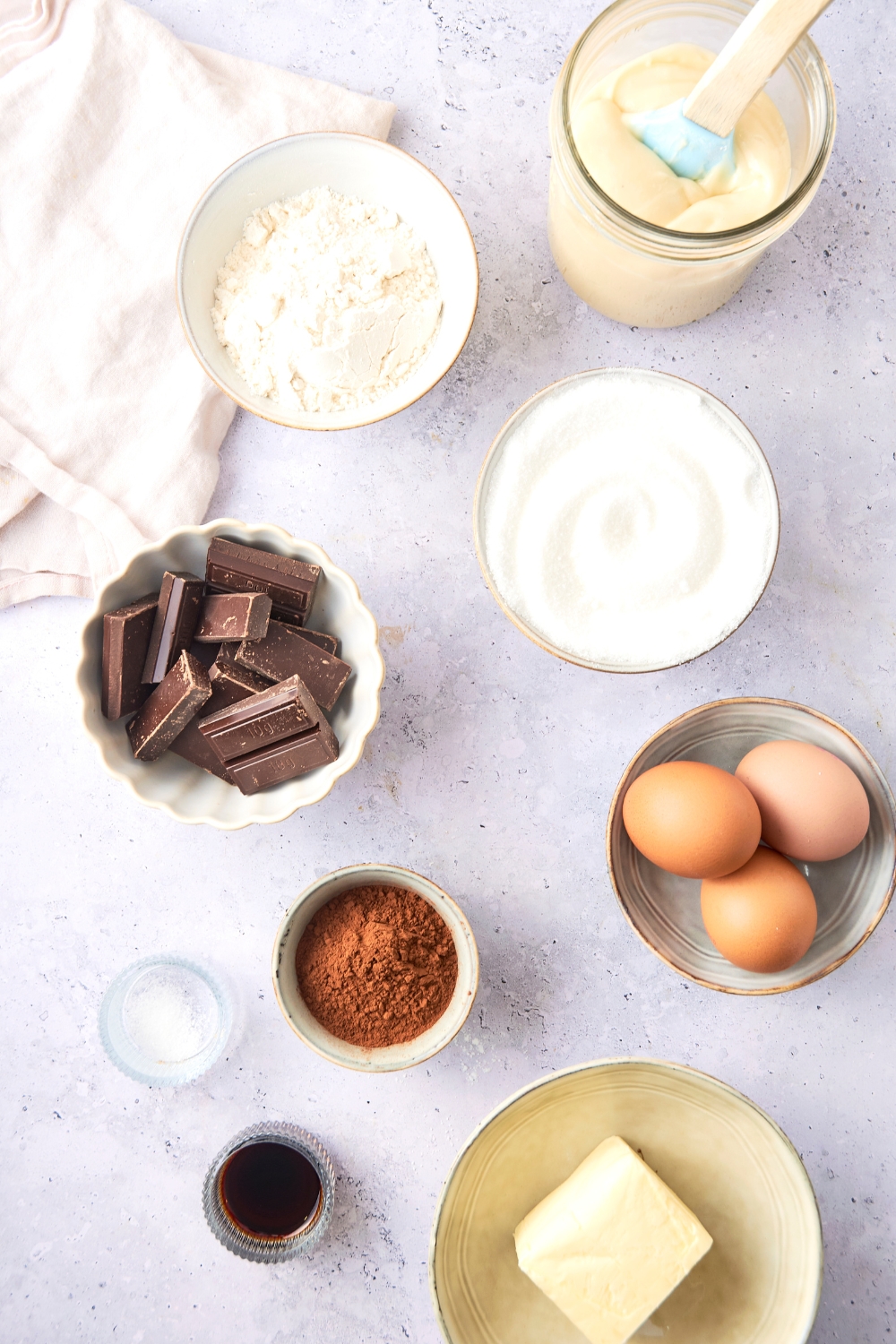 A countertop with various bowls containing cream cheese, flour, chocolate, eggs, butter, cocoa powder, vanilla, salt.