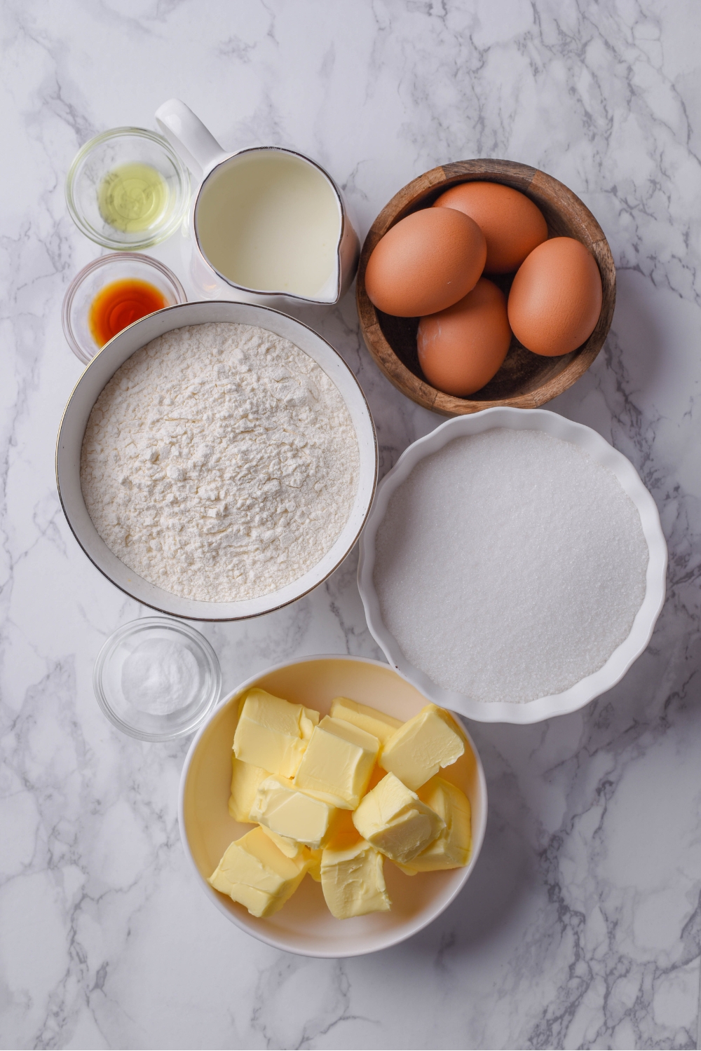 Eggs, flour, sugar, butter, buttermilk, vanilla, and salt are all in separate containers on a gray counter top.