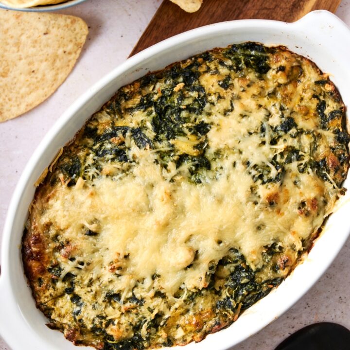 A casserole dish with baked Cheddar's spinach dip on a wooden serving board. Tortilla chips are next to the board.