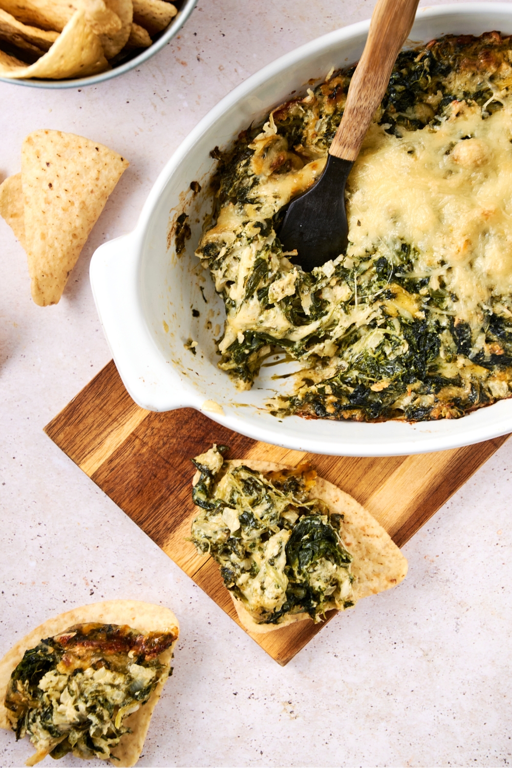 A casserole dish with baked Cheddar's spinach dip on a wooden serving board. Tortilla chips are next to the board. Two tortilla chips have heaping scoops of the spinach dip on it.