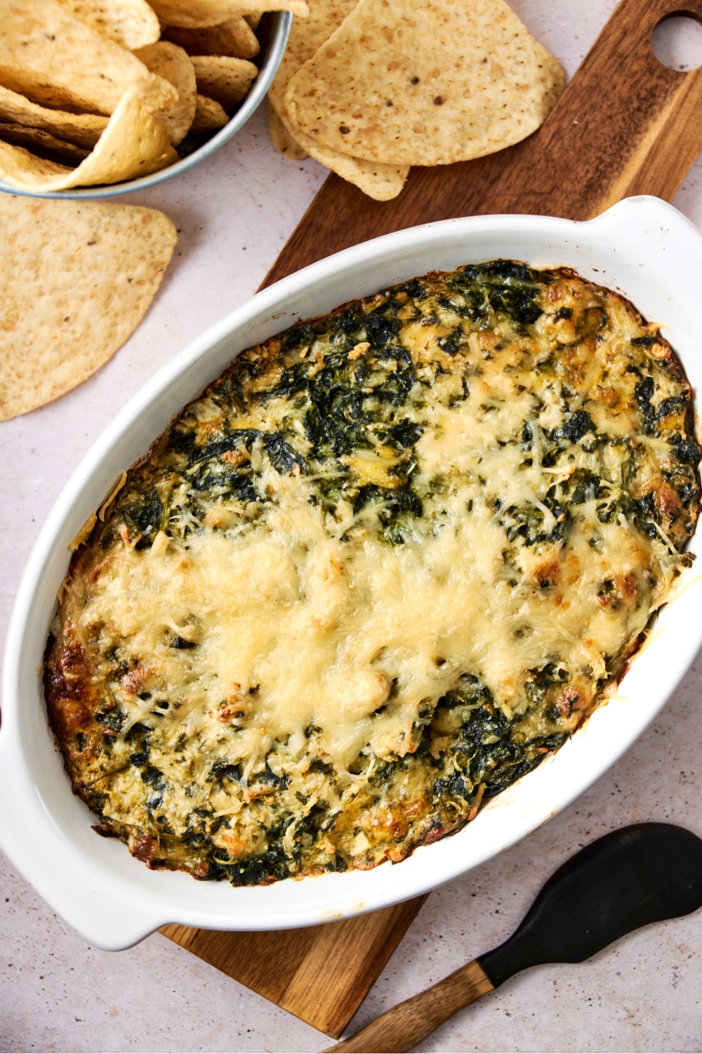 A casserole dish with baked Cheddar's spinach dip on a wooden serving board. Tortilla chips are next to the board.