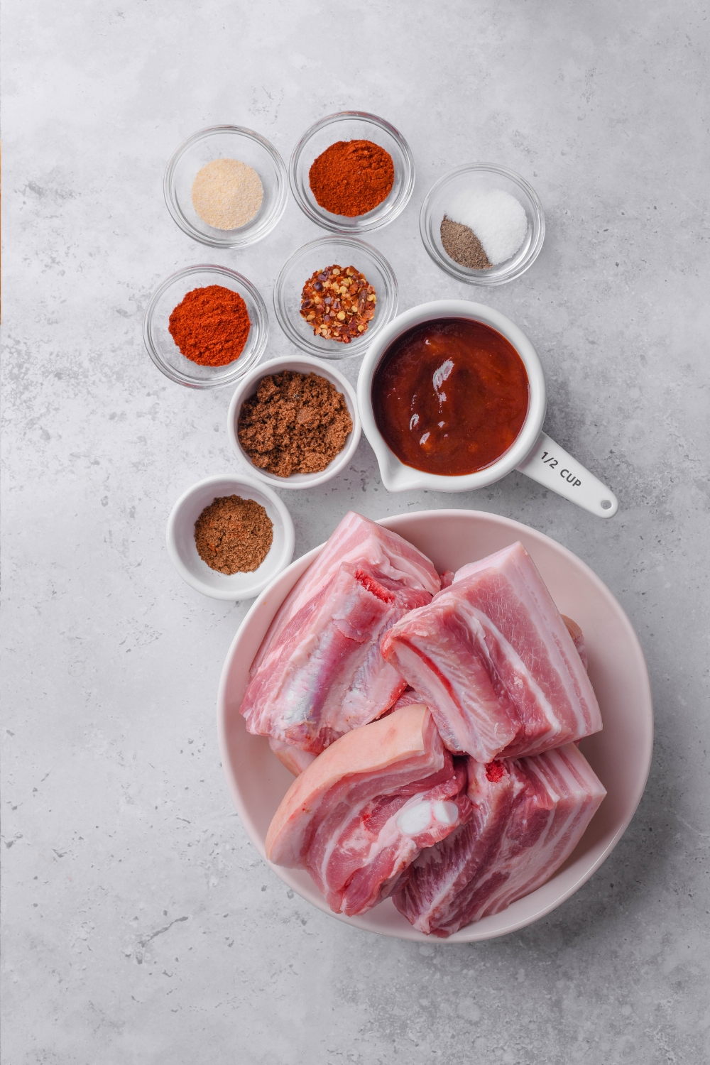 Smoked paprika, onion powder, cumin, salt, pepper, barbecue sauce, brown sugar, and red pepper flakes are all in separate glass bowls on a gray counter top. A large white plate is nearby with raw country style ribs on it.