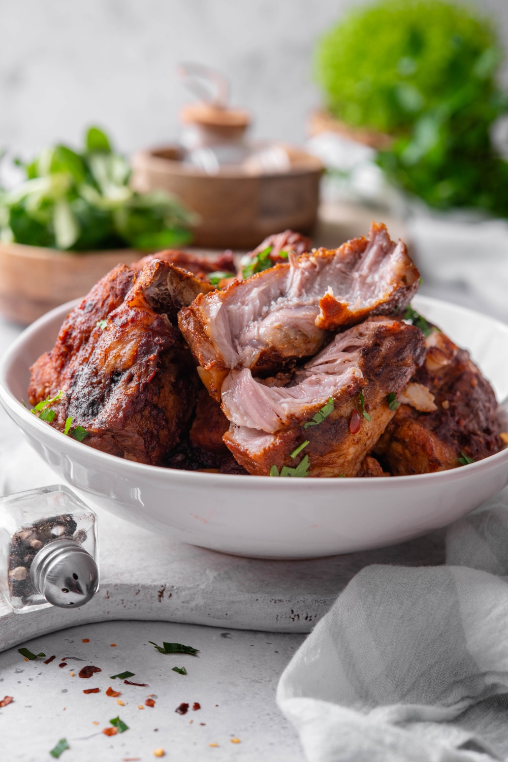 A white bowl is full of country style ribs. The ribs are perfectly cooked. One of the ribs is cut in half and they are garnished with parsley.