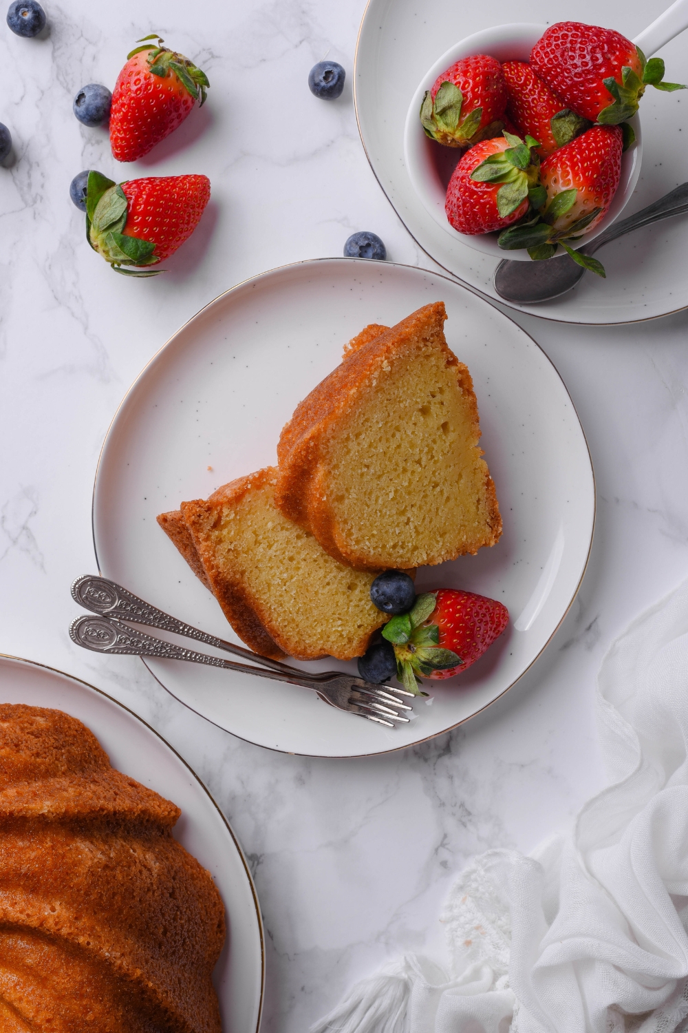 Two slices of buttermilk pound cake are sitting on a white plate. Fresh berries are scattered nearby and two forks rest on the plate.