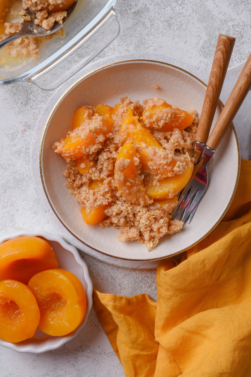 A white bowl holds peach crisp with canned peaches. Two forks rest in the bowl. A small bowl nearby holds extra canned peaches.