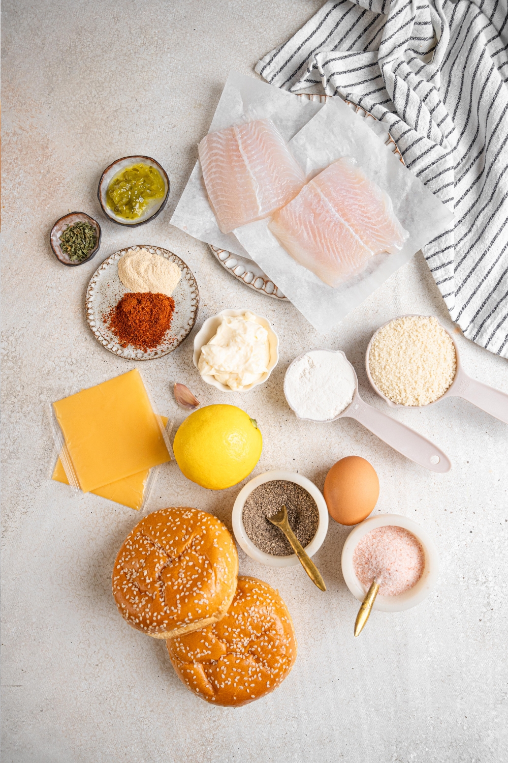 Burger buns, salt, pepper, a lemon, cheddar cheese, flour, mayonnaise, panko, seasonings, and fish filets sit in separate containers on a gray counter top.