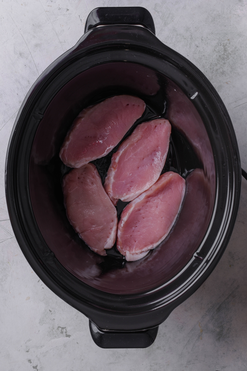 A black crock pot holds four raw pork chops.