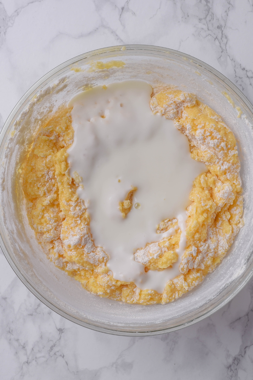 A glass bowl holds buttermilk pound cake batter. Buttermilk is sitting on the batter ready to be mixed in.