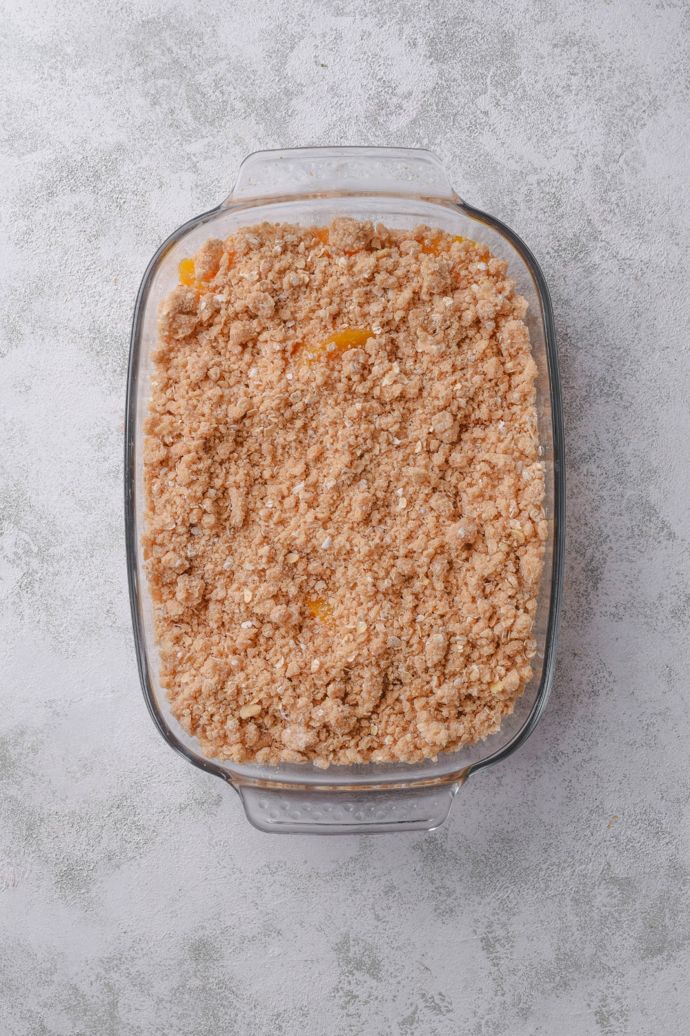A glass baking dish holds an assembled peach crisp with canned peaches.