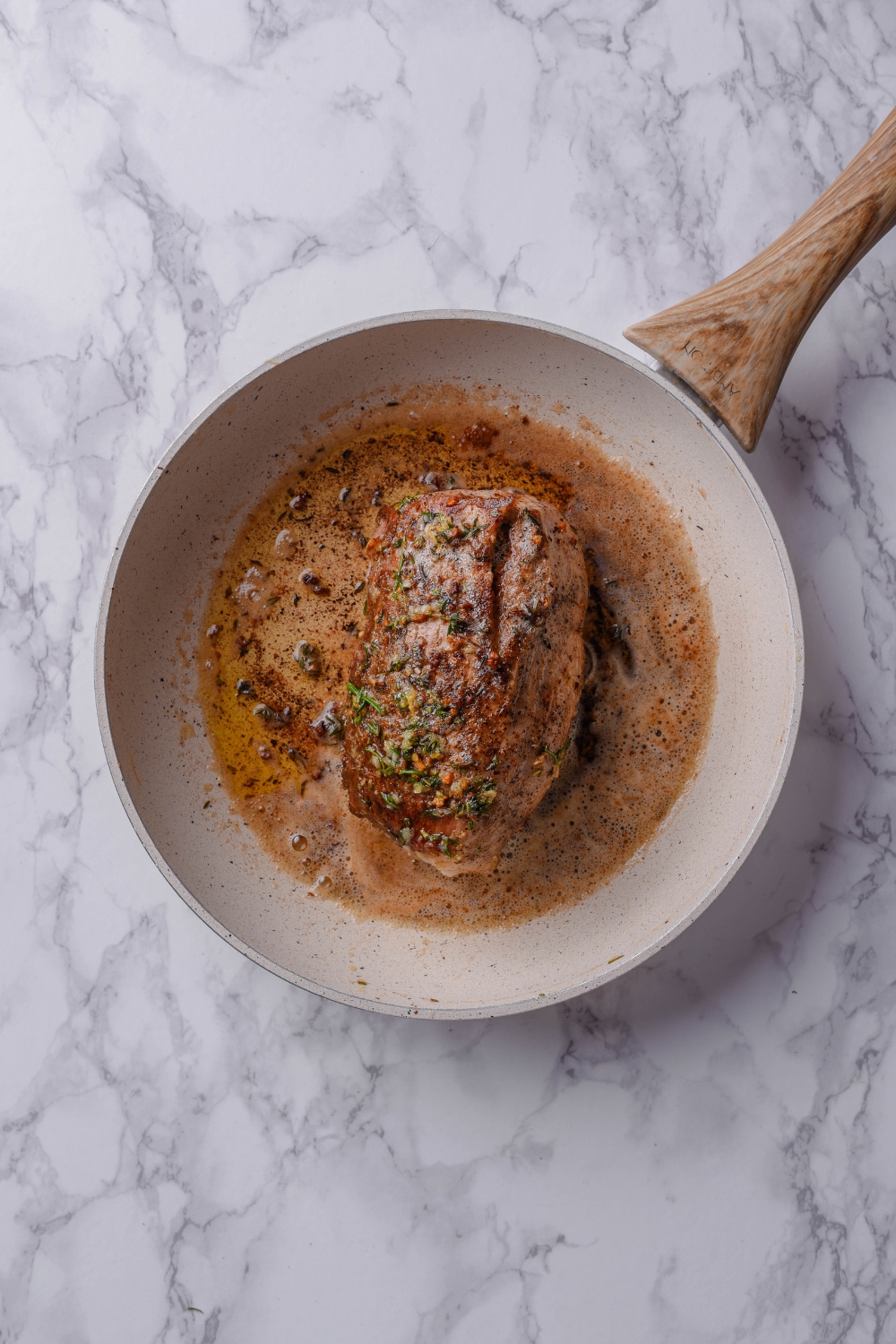 A white frying pan holds a perfectly cooked venison tenderloin and lots of melted herb butter.