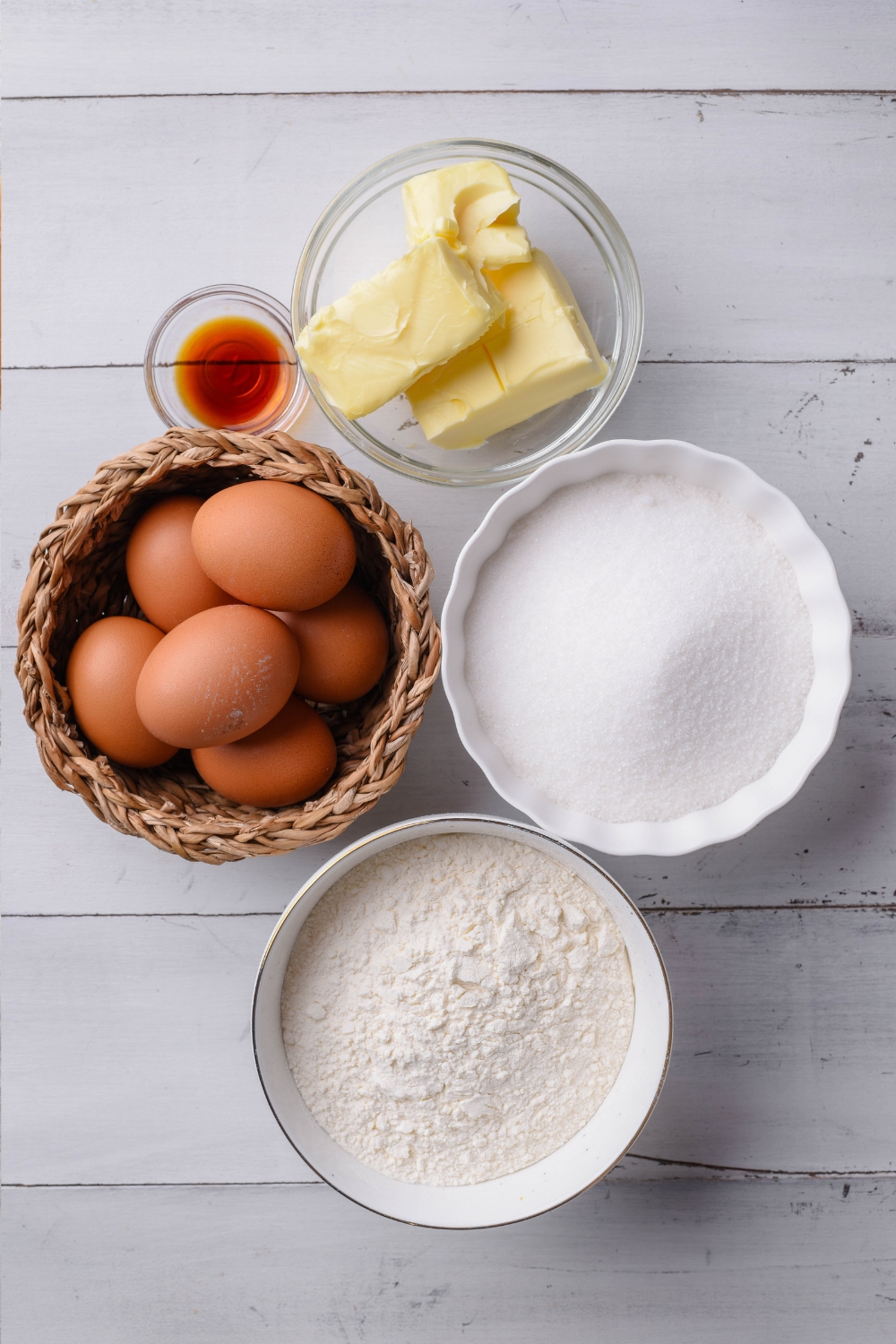 Butter, eggs, flour, sugar, and vanilla are in separate bowls on a gray counter top.