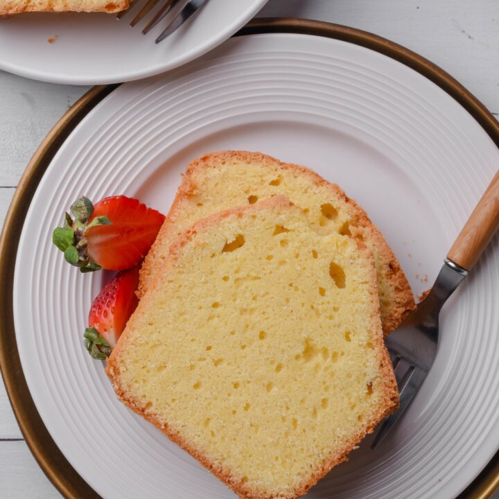 A white plate holds two thick slices of old fashioned pound cake. A fork and two strawberries are on the plate.