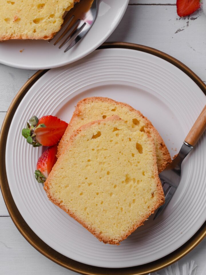 A white plate holds two thick slices of old fashioned pound cake. A fork and two strawberries are on the plate.