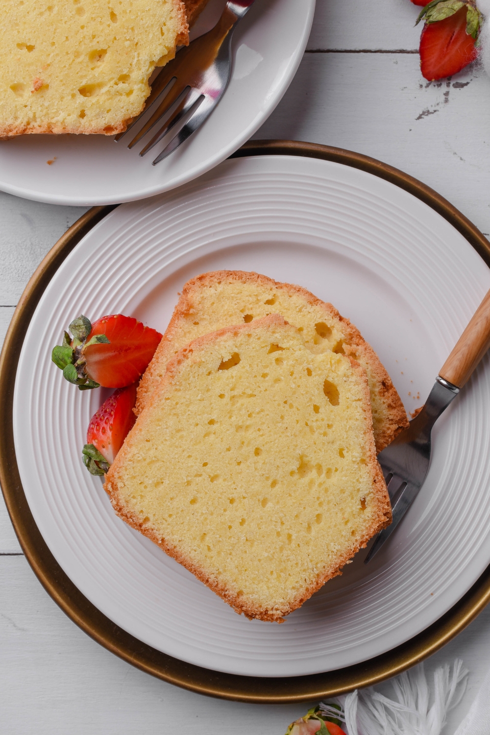 A white plate holds two thick slices of old fashioned pound cake. A fork and two strawberries are on the plate.