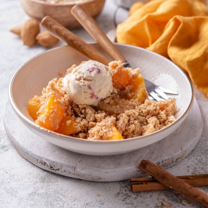 A white bowl holds a large serving of peach crisp with canned peaches. The crisp is topped with a scoop of ice cream and a fork rests in the bowl.