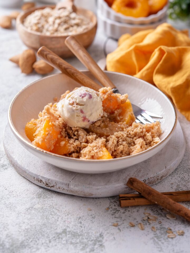 A white bowl holds a large serving of peach crisp with canned peaches. The crisp is topped with a scoop of ice cream and a fork rests in the bowl.