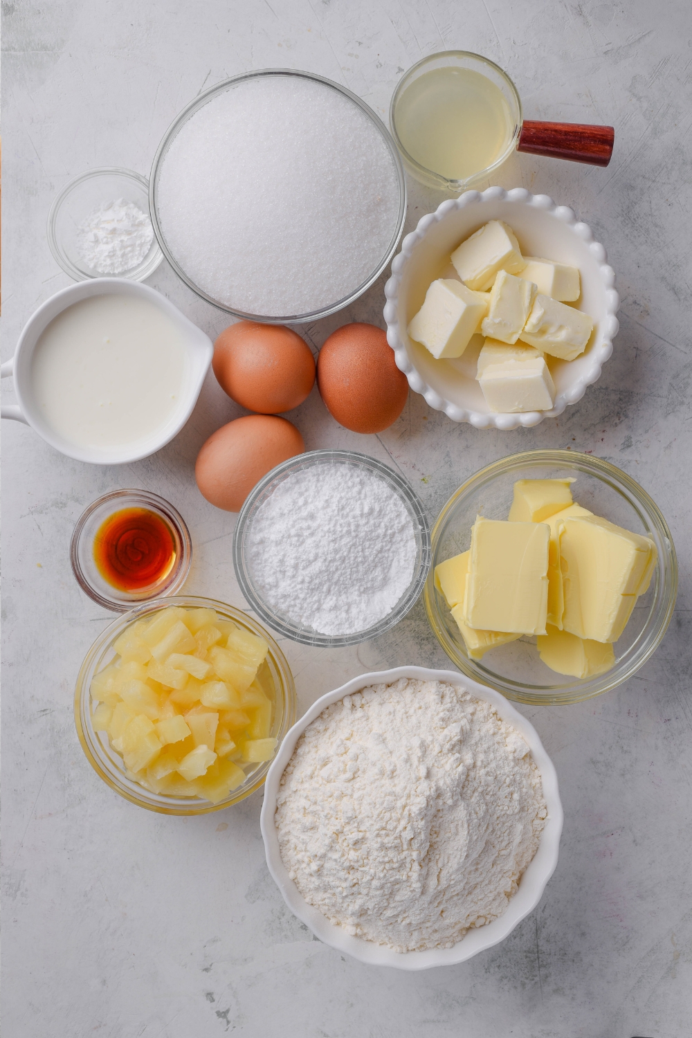Flour, pineapple, butter, powdered sugar, vanilla, vegetable shortening, eggs, buttermilk, sugar, and pineapple juice are all in separate bowls on a counter top.