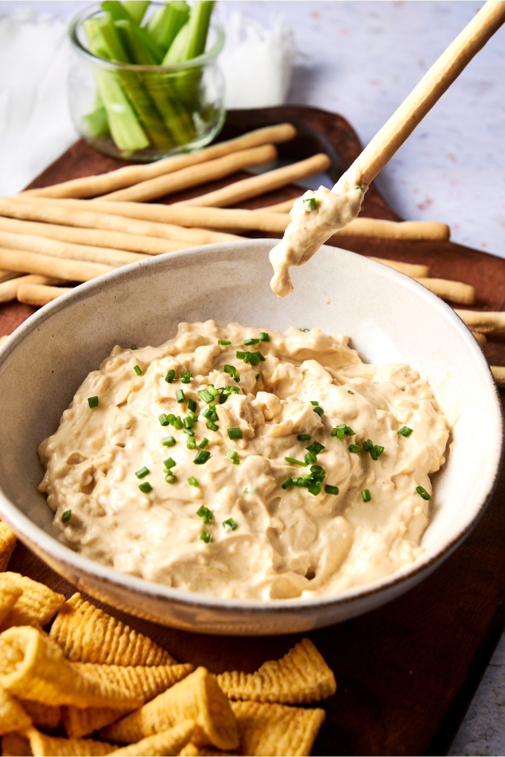 A large white bowl is full of Lipton french onion dip garnished with chives. Someone scoops up the dip with a breadstick.