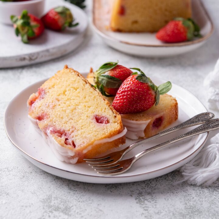 Two slices of strawberry pound cake are on a white plate. There are fresh strawberries and forks on the plate also.