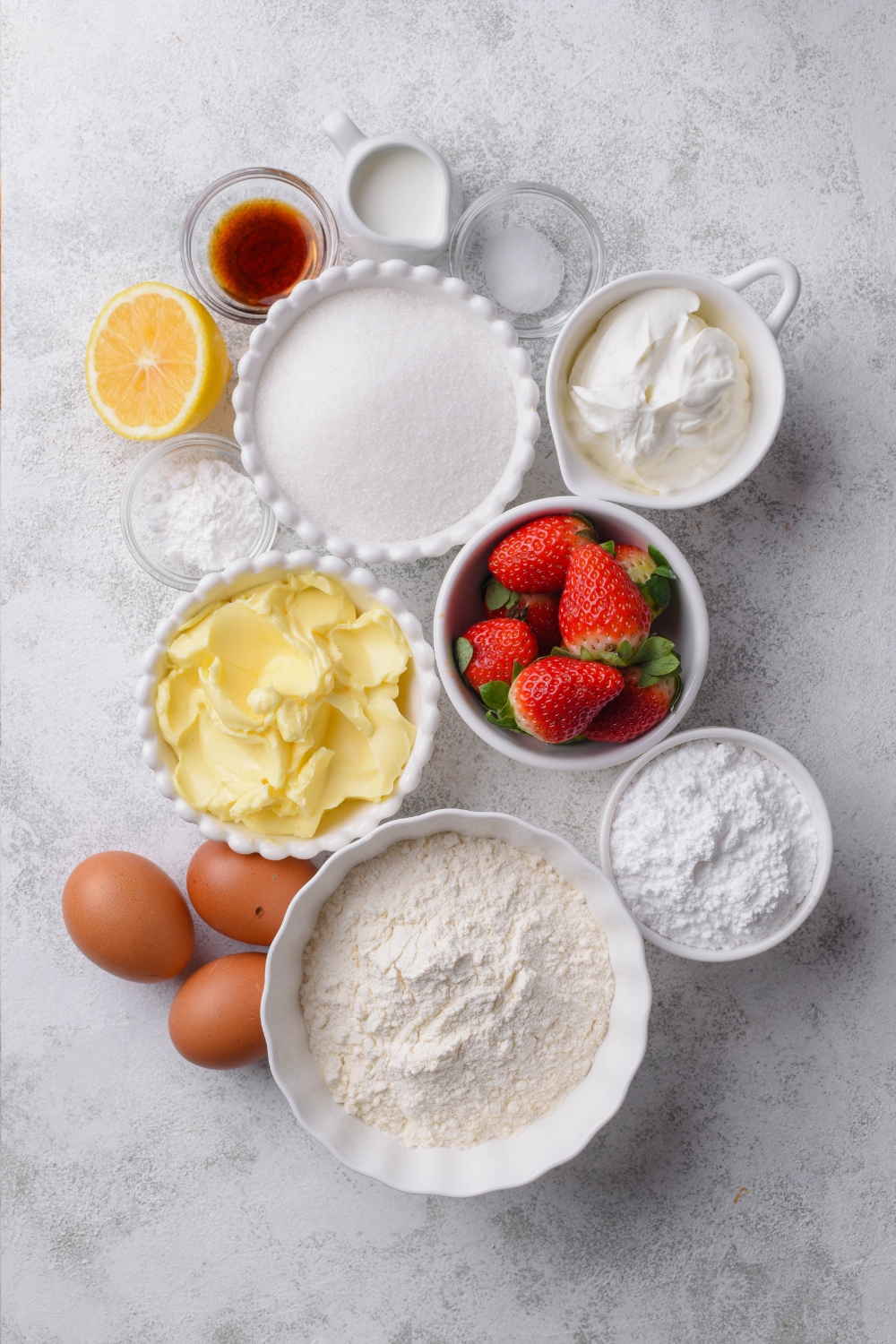 Flour, eggs, baking powder, baking soda, butter, strawberries, icing sugar, half of a lemon, sour cream, salt, and milk are all in separate containers on a gray counter top.