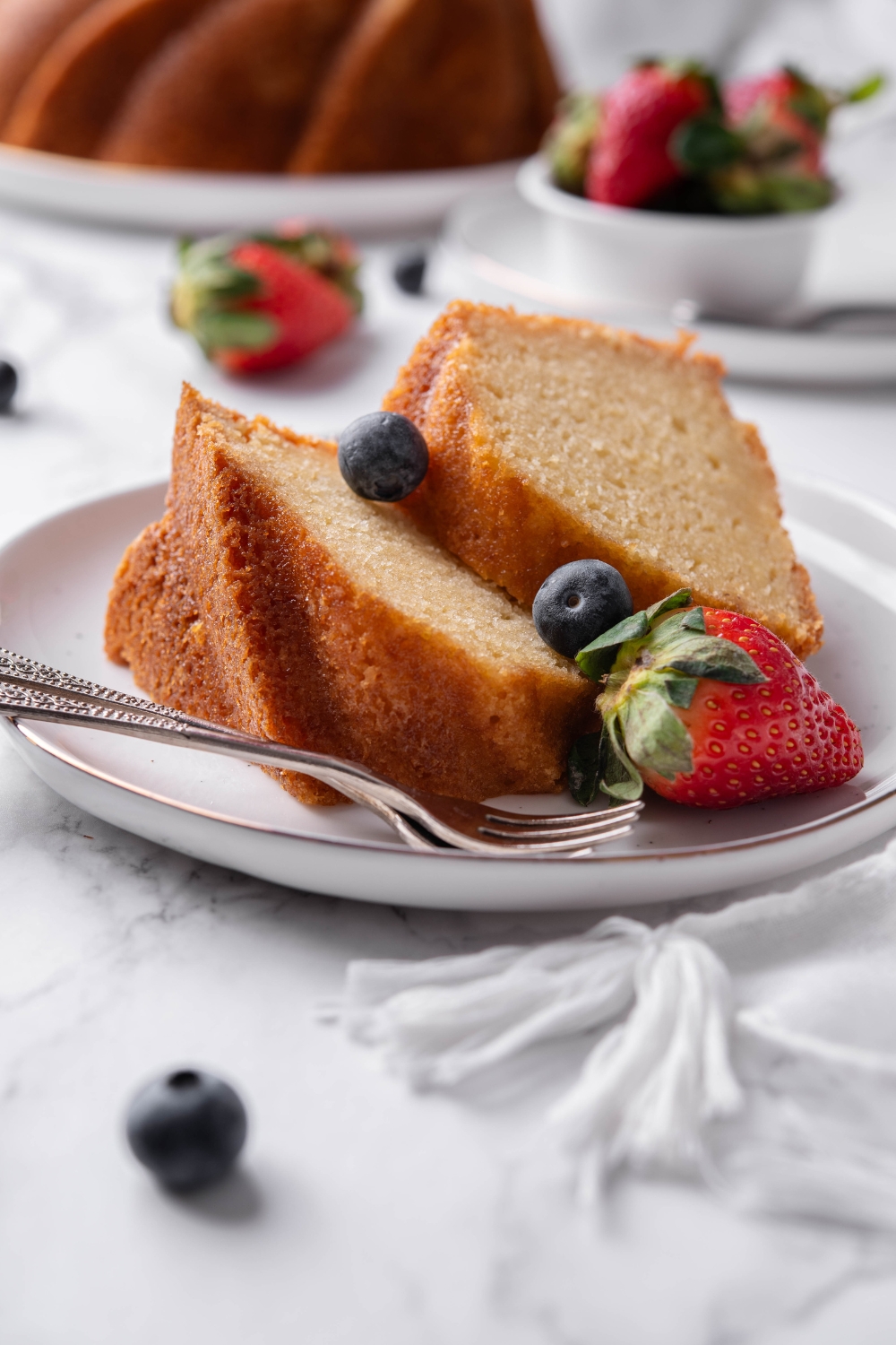 Two slices of buttermilk pound cake sit on a white plate. There are fresh berries and two forks resting on the plate as well.