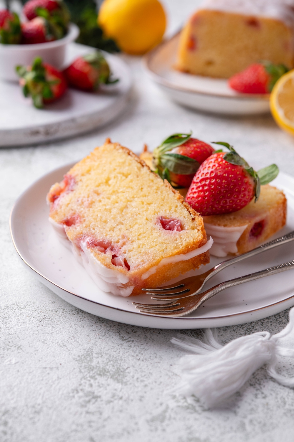 Two slices of strawberry pound cake sit on a white plate. There are two fresh strawberries on the plate as well.