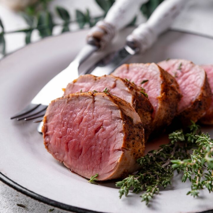A white plate holds a sliced venison tenderloin. A fork and knife are on the plate, as well.