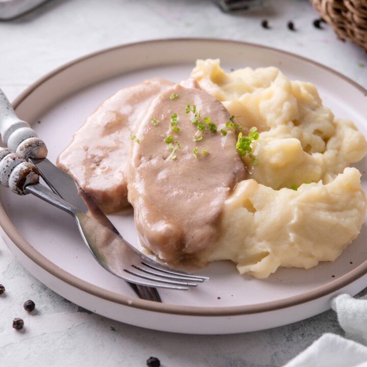 Two pork chops covered in a gravy on a white plate with mashed potatoes.