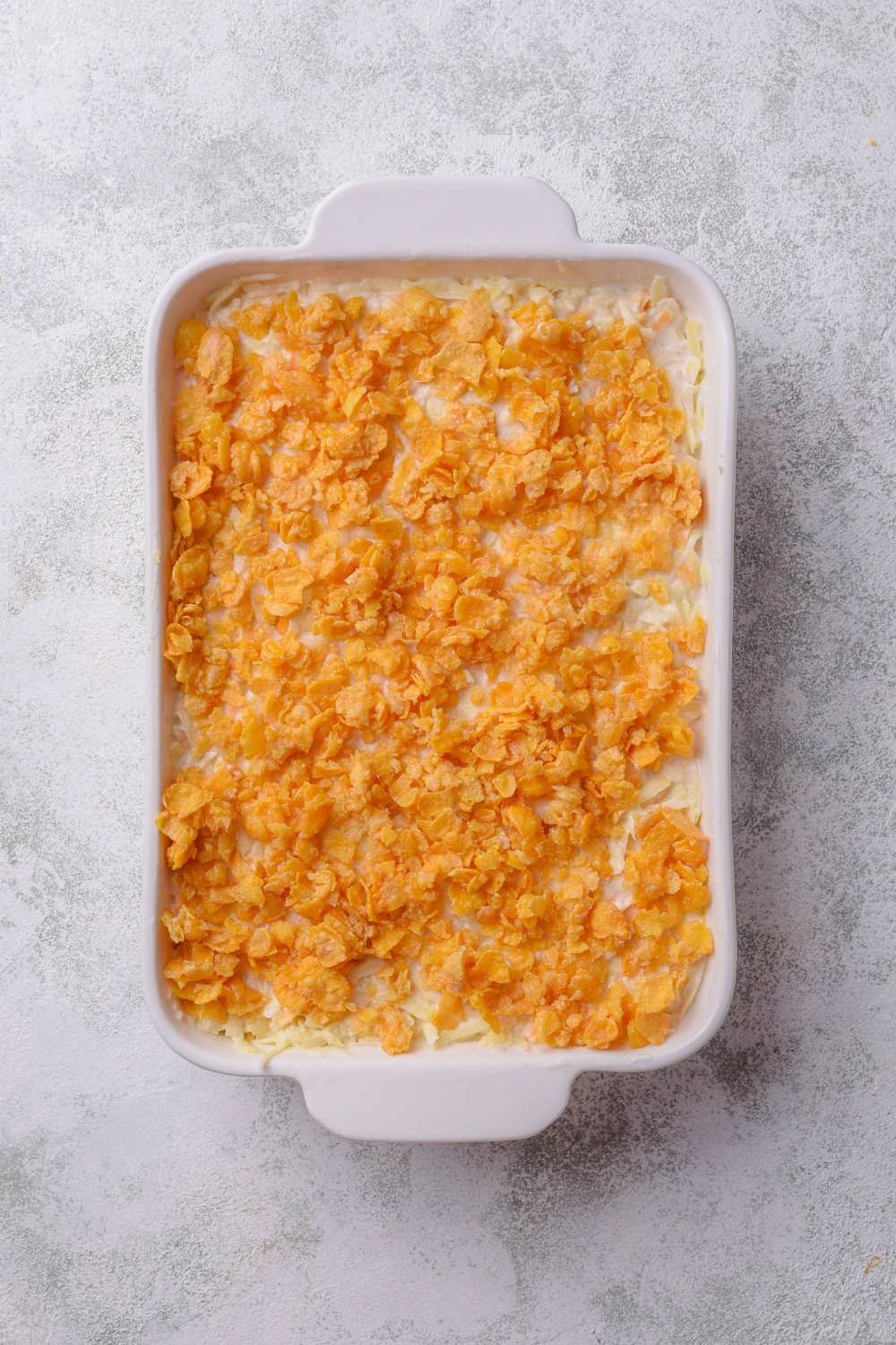 A casserole dish with unbaked hash brown casserole topped with crushed corn flakes.