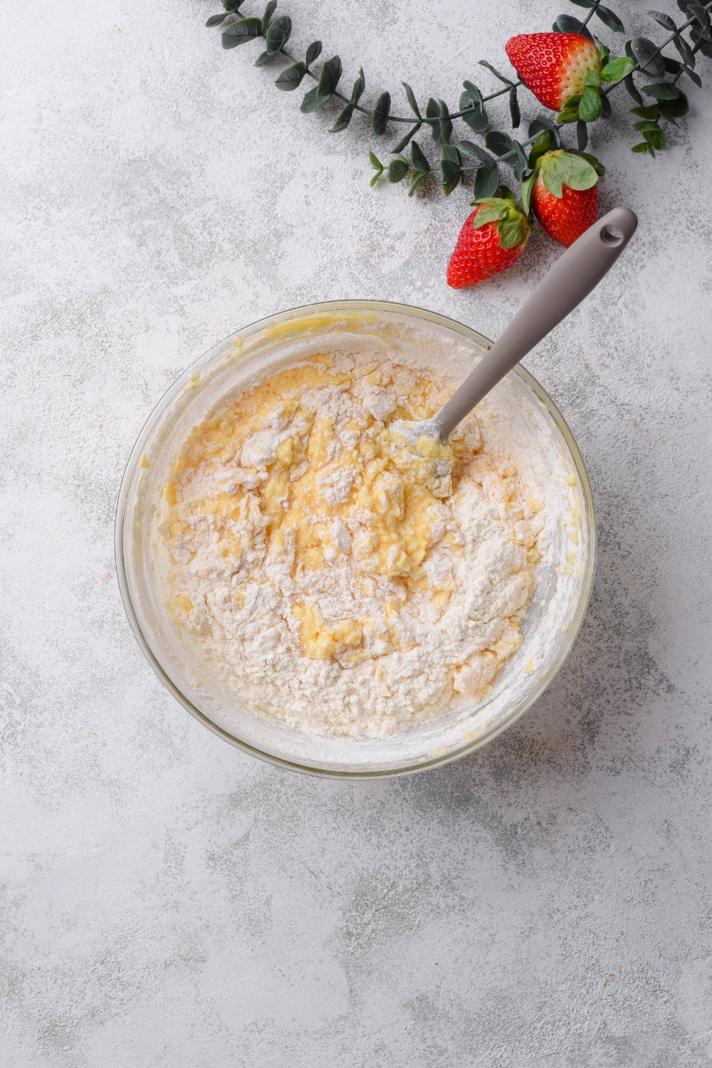 A glass bowl holds cake batter and flour. The flour has begun to be mixed in. A spoon rests in the bowl as well.