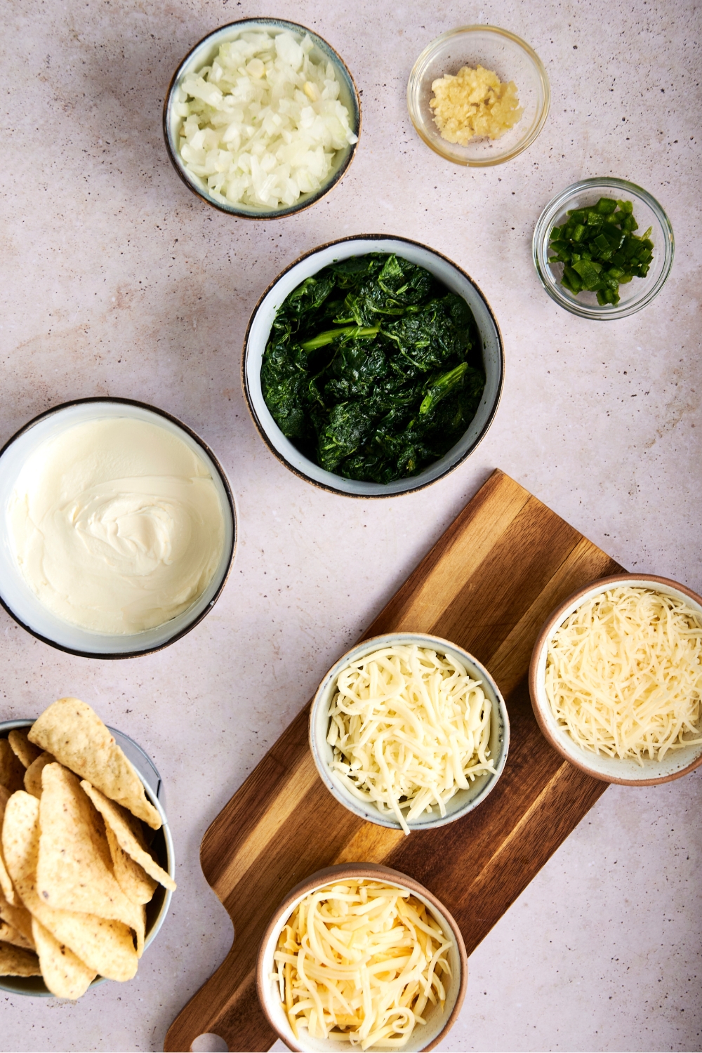 A countertop with multiple bowls with measured out portions of spinach, cream cheese, diced onion, jalapeno, minced garlic, three shredded cheeses, and tortilla chips.
