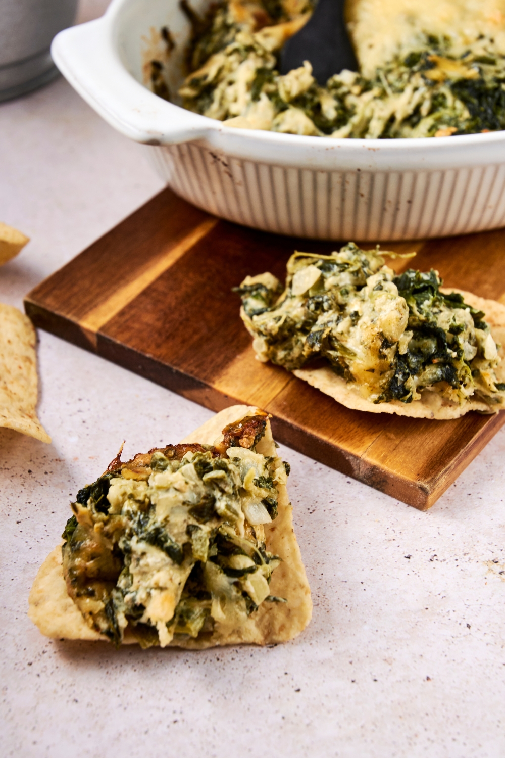 A casserole dish with baked Cheddar's spinach dip on a wooden serving board. Tortilla chips are next to the board. Two tortilla chips have heaping scoops of the spinach dip on it.