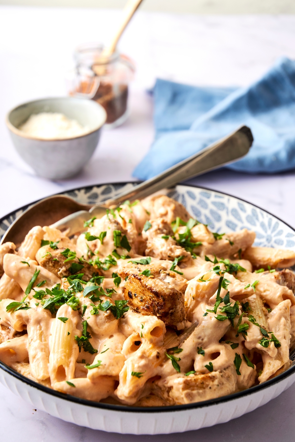 A blue and white serving bowl is piled high with Buffalo wild wings garlic parmesan chicken pasta. The dish has been garnished with fresh herbs. A set of serving utensils rest in the bowl, as well.