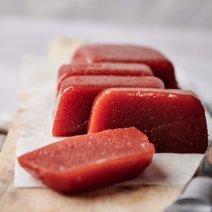 A sliced block of guava paste is resting on a wooden serving board. The board is lined with parchment paper and a knife rests on the board, also.