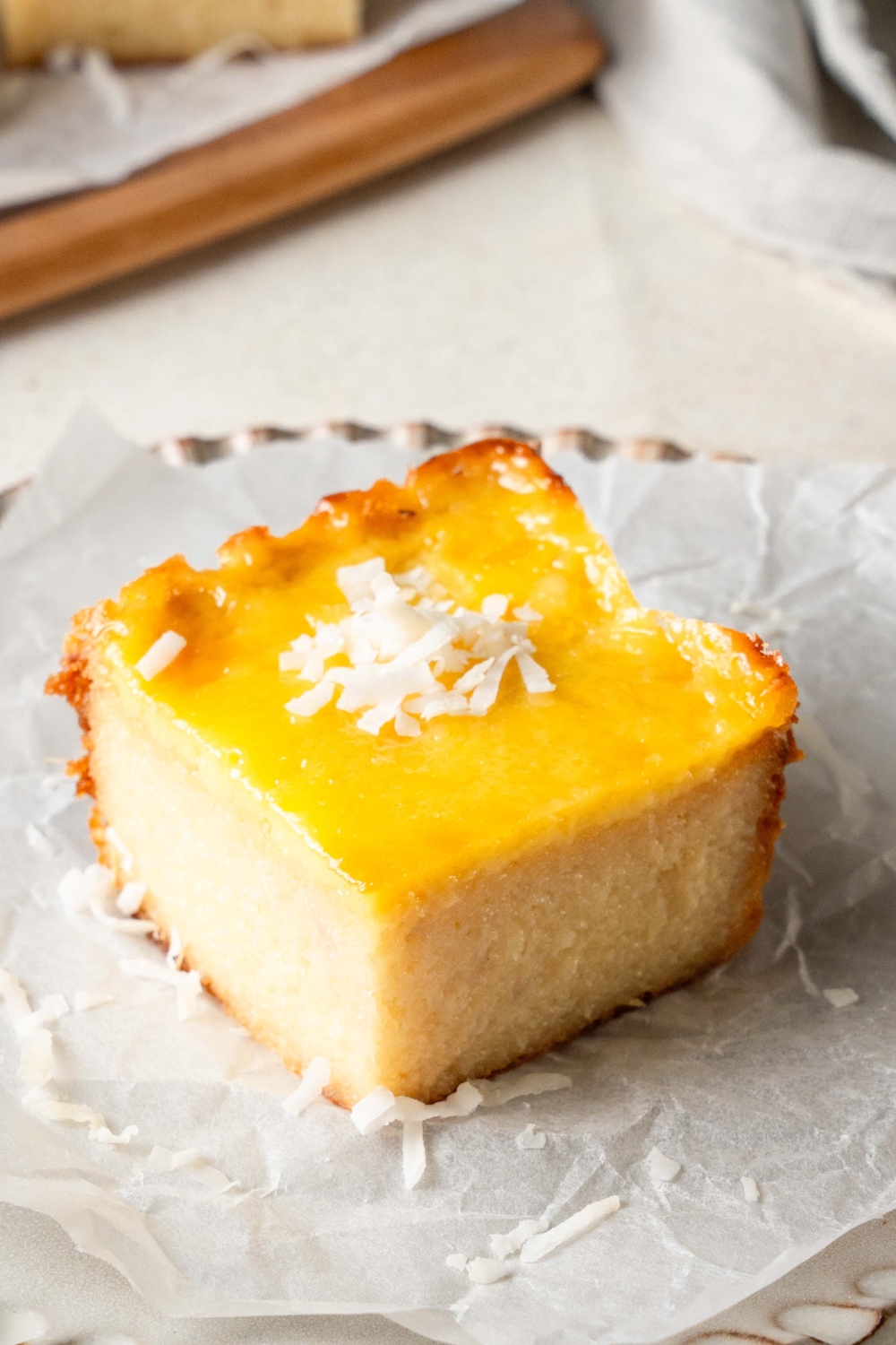 A square piece of cassava cake sits on a plate lined with parchment paper. The piece of cake is topped with shredded coconut.